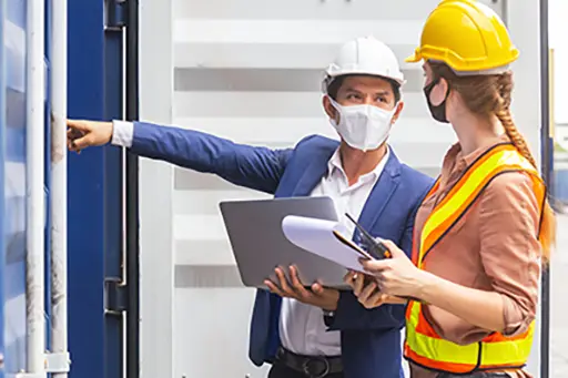 Teamwork foreman man and woman wearing protection face mask and safety helmet using laptop and holding clipboard checking containers in cargo ship for import export, Industrial container cargo concept.